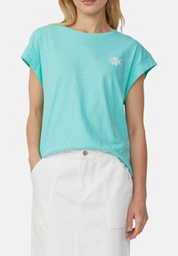 Vrouw in een losse turquoise top met korte mouwen en een klein wit visontwerp, gecombineerd met een hooggerote witte rok met zakken.