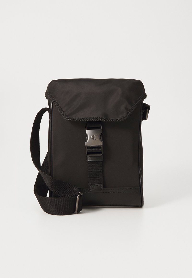 Bolso cruzado negro de nailon con solapa plegable, correa ajustable y hebilla de hardware plateado. Textura suave, diseño minimalista.