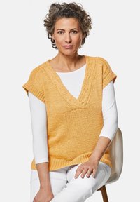 Gebreid mouwloos topje in fel geel-oranje met een V-hals, een gemêleerd patroon en ribbelrand, gelaagd over een wit longsleeve shirt.