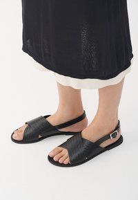 Μαύρες πλεκτές δερμάτινες σανδάλια με ανοιχτό μπροστινό μέρος, σταυρωτές λουρίδες και λουρί με πόρπη στον αστράγαλο. Επίπεδη σόλα χωρίς σημαντικά διακοσμητικά στοιχεία.