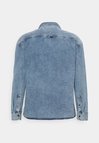 Ljusblå denimjacka sedd bakifrån, med långa ärmar, knappade manschetter och en liten röd, vit och blå etikett nära kragen och manschetterna.