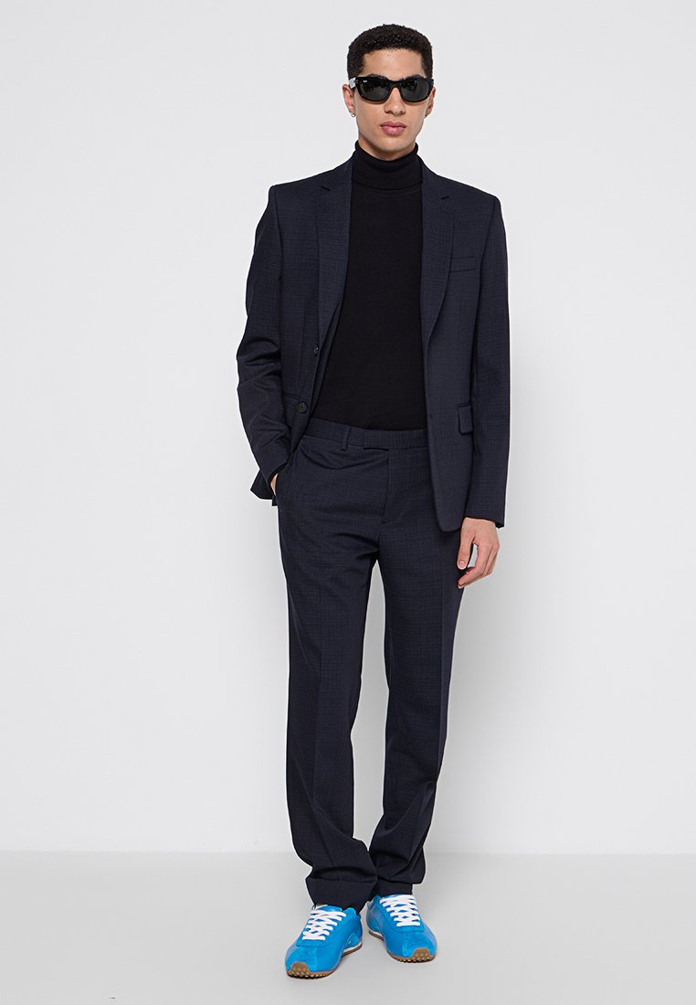 Traje sastre azul marino con un acabado texturizado, combinado con un cuello alto negro. Zapatillas deportivas azules, gafas de sol negras y un diseño ajustado.