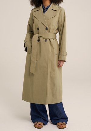 Nainen pukeutuneena beigenväriseen kaksiriviseen trenssitakkiin, sinisiin farkkuihin ja vaaleanruskeisiin avojalkaisiin sandaaleihin, seisoo yksinkertaisen taustan edessä.