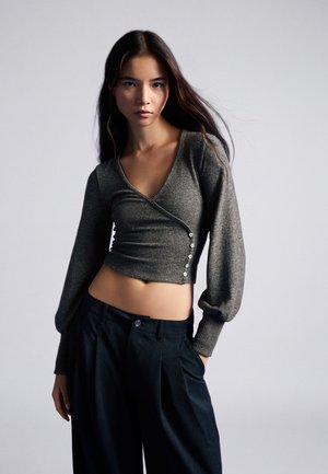Junge Frau mit langen dunklen Haaren, die einen grauen geknöpften Wickelpullover und lockere schwarze Hosen trägt, posiert vor einem einfarbigen Hintergrund.