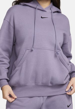 Kvinde iført en lavendel Nike hættetrøje med frontlomme og matchende lavendel joggingbukser, hænder holder i kanten af hættetrøjen.