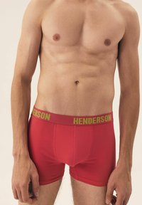 Czerwone slipy z zielonym paskiem z napisem "HENDERSON". Wykonane z miękkiej tkaniny, charakteryzujące się dopasowanym krojem i gładką fakturą.