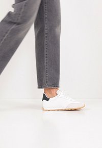 Λευκό αθλητικό παπούτσι με υφή στη μύτη, μαύρη διακόσμηση στη φτέρνα και καφέ σόλα, σε συνδυασμό με γκρι τζιν με ίσια γραμμή.