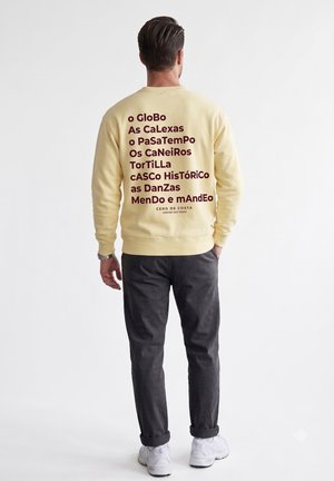 Hombre con sudadera amarillo pálido con texto estilizado en rojo oscuro en la espalda, pantalones oscuros y zapatillas blancas, de pie sobre fondo blanco.