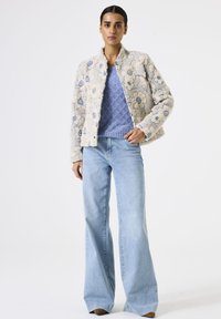 Frau in einer cremefarbenen, blumig strukturierten Jacke über einem blauen Strickpullover, mit hellblauen, weit geschnittenen Jeans, die mit einer Hand in der Tasche steht.