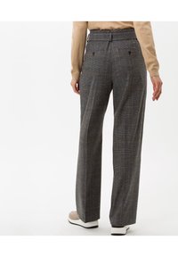 Pantalon à carreaux gris avec taille haute, jambes larges et tissu texturé. Comprend des poches arrière avec fermetures à boutons. Associé à des chaussures beige.