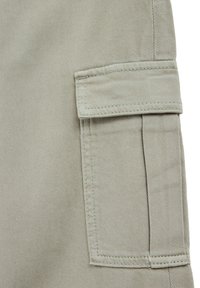 Gai bēšas kravas bikses ar teksturētu virsmu, kurām ir sānu kabata ar vāku un nostiprināta šuves gar malām.