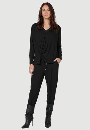 Femme aux longs cheveux foncés portant un haut drapé à manches longues noir, un pantalon noir à rayures et des bottes à talons noirs sur un fond clair.
