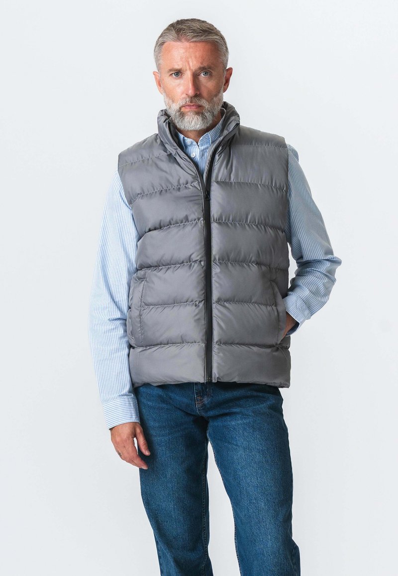 Grå polstret vest med høj krave, lynlås foran og vandret syninger. Båret over en blå og hvidstribet skjorte og kombineret med blå jeans.