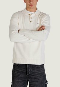 Biały dziany sweter z kołnierzem polo, trzema brązowymi guzikami, prążkowanymi mankietami i dołem, oraz małym logo marki na piersi. Noszony z ciemnymi jeansami.