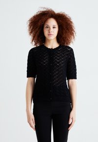 Cardigan noir en tricot à manches courtes, avec un motif en losanges, des bords côtelés et une fermeture à boutons. La texture semble légère et aérée.