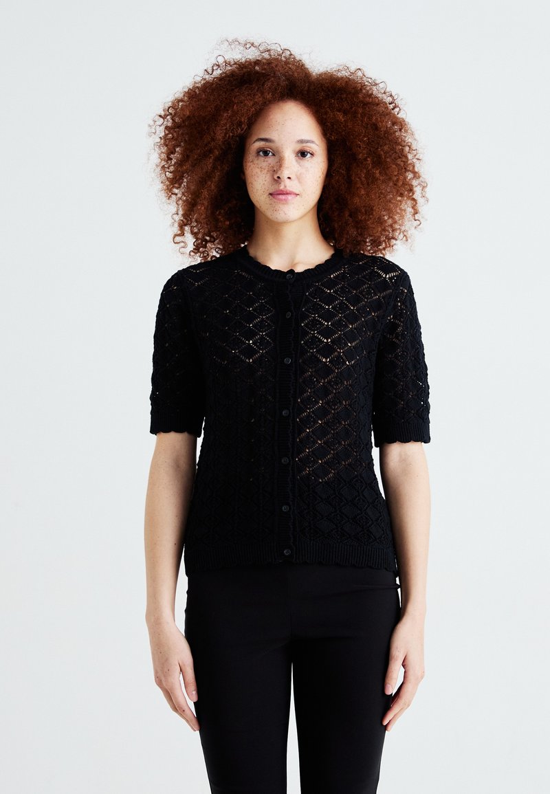 Cardigan noir en tricot à manches courtes, avec un motif en losanges, des bords côtelés et une fermeture à boutons. La texture semble légère et aérée.