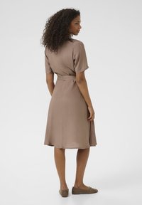Robe brune longueur genou en tissu léger, manches courtes, taille cintrée et jupe évasée ; assortie à des chaussures plates taupe.