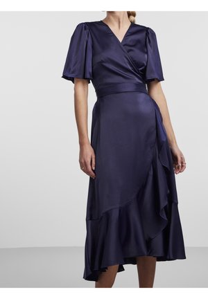 Kobieta w ciemnoniebieskiej satynowej sukience kopertowej z krótkimi bufkami i asymetrycznym falbanowanym dołem na jednolitym tle.