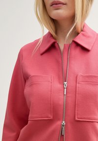 Veste rose zippée en tissu texturé avec un col, comportant deux poches avant et une fermeture éclair argentée.