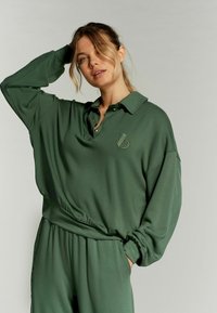 Polo sweatshirt oversized verde scuro con colletto e logo ricamato, caratterizzato da una vestibilità comoda e un elastico in vita. Tessuto morbido.