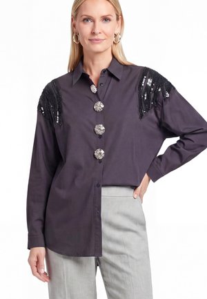 Donna che indossa una camicia scura a maniche lunghe con grandi bottoni decorativi e frange nere di paillettes sulle spalle, abbinata a pantaloni grigio chiaro.