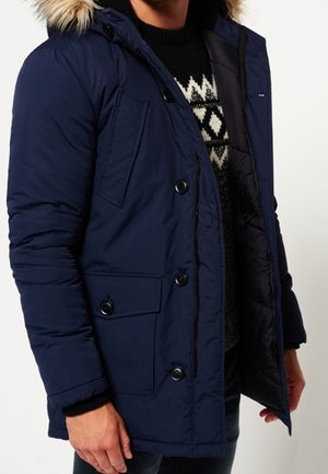 Homme portant un manteau d'hiver bleu marine avec une capuche bordée de fourrure, un pull à motifs en dessous, debout devant un fond uni.