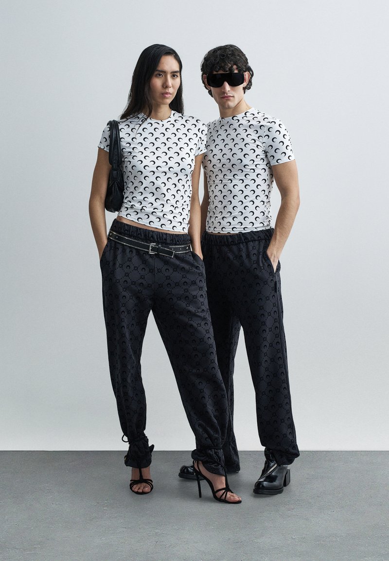 T-shirts brancas de manga curta com design preto em forma de crescente, combinadas com calças de jogging pretas com padrão. Os acessórios incluem uma mala preta e óculos de sol de tamanho grande.