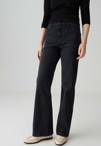 Vrouw in hooggetailleerde zwarte rechte broek, zwart langemouwen top en crèmekleurige loafers, staand tegen een effen lichte achtergrond.