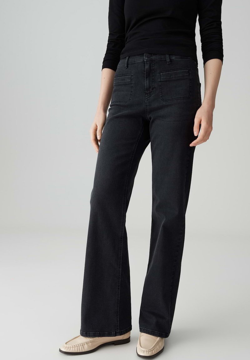 Vrouw in hooggetailleerde zwarte rechte broek, zwart langemouwen top en crèmekleurige loafers, staand tegen een effen lichte achtergrond.