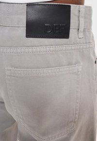 Grey denim pants featuring a black leather waistband patch with embossed lettering, back pocket with subtle stitching detail.