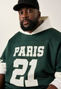 Camiseta deportiva verde con acentos blancos, que presenta "PARIS" y "21" impresos en estilo perforado. Se ve el cuello con capucha debajo. Lleva una gorra negra.
