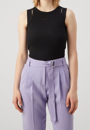Kobieta ubrana w dopasowany czarny top bez rękawów z logo "BOSS" oraz wysokowalne liliowe spodnie z zakładkami i materiałowym paskiem z metalową klamrą.