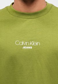 Maslinasto zeleni pamučni T-shirt s okruglim izrezom, s bijelim printom "Calvin Klein JEANS" na prednjoj strani. Glatka tekstura.