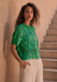 Bluza verde din dantelă cu decupaje florale, cu nasturi aurii și mâneci scurte. Pantaloni bej deschis, plisati, cu buzunare laterale.