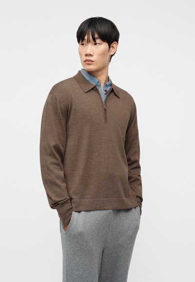 Νέος άνδρας που φοράει καφέ πουλόβερ με φερμουάρ πάνω από μπλε πουκάμισο με γιακά και γκρι παντελόνι, στέκεται με τα χέρια στις τσέπες, κοιτώντας αριστερά, με λευκό φόντο.