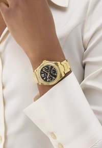 Gold watch with a black dial, two subdials, crystal accents on the bezel, and a gold metal bracelet, worn on a wrist against a light-colored shirt.