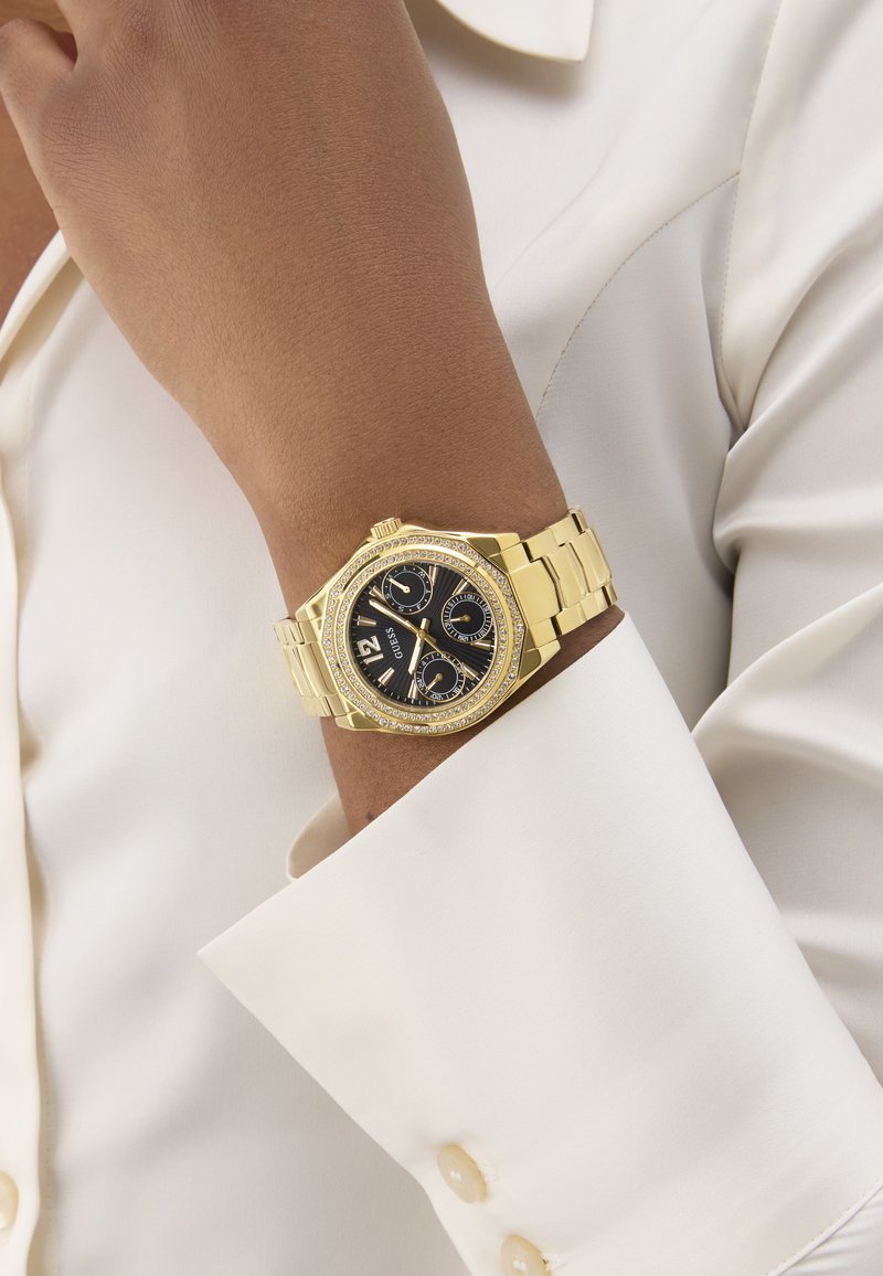 Gold watch with a black dial, two subdials, crystal accents on the bezel, and a gold metal bracelet, worn on a wrist against a light-colored shirt.