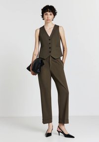 Olivgrüne Weste mit fünf schwarzen Knöpfen, kombiniert mit passenden taillierten Hosen und schwarzen spitzen Slingback-Absätzen. Schwarze Clutch in der Hand.
