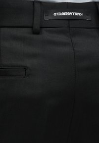 Κοντινό πλάνο μαύρου παντελονιού με ραφές που δείχνει τη μέση, τις θηλιές της ζώνης, την πίσω τσέπη και μια ανάποδη ετικέτα της μάρκας Karl Lagerfeld.