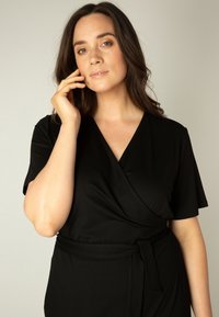 Zwarte wikkeljurk met korte mouwen, een soepele textuur en een ceintuur in de taille. Heeft een V-hals en een getailleerd silhouet.