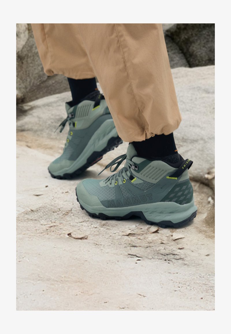 Scarpe da esterno Mammut in verde spento con tomaia testurizzata, dettagli gialli e suola nera robusta. Design durevole per le attività all'aperto.