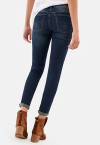 Jeans ajustados de denim azul oscuro, con una cintura de altura media, dos bolsillos traseros y bajos remangados, combinados con botas de tobillo de tacón marrones.