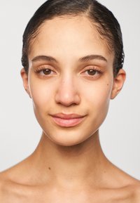 Gros plan sur un visage avec une peau lisse, des yeux brun clair et des cheveux sombres et mouillés. L'arrière-plan est gris uni, mettant en valeur les traits et les contours du visage.