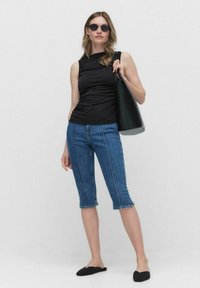 Top noir sans manches, jean court bleu avec coutures latérales, chaussures plates noires, sac fourre-tout noir et lunettes de soleil rondes sur un fond uni.