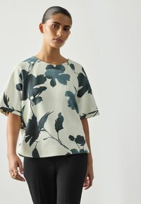 Kvinna med släta hår som bär en kortärmad krämfärgad topp med stor svart blomstertryck och svarta byxor mot en enfärgad ljus bakgrund.