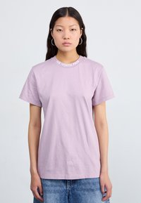 Camiseta de algodón color púrpura claro, de manga corta, cuello redondo con el logotipo "THE NORTH FACE" bordado en blanco alrededor del cuello. Ajuste casual.