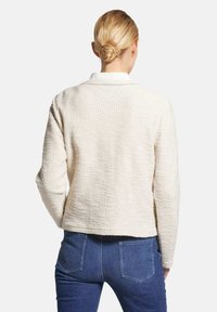 Cremfarbener, strukturierter Blazer mit langen Ärmeln, einem Kragen und einem cropped Design, kombiniert mit dunkelblauen Jeans aus Denim mit Gesäßtaschen.