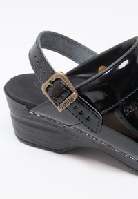 Zwarte gepatineerde leren sandaal met een chunky hak, verstelbare band en een metalen gesp. Voorzien van een getextureerd plateau en gestikte details.