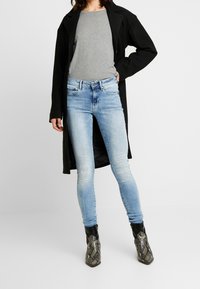 Person wearing light blue skinny jeans, gray top, black long coat, and snake-print ankle boots, standing against a white background.