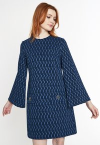 Navyblaues Kleid mit langen Glockenärmeln, strukturiertem Stoff mit geometrischem Muster und zwei Fronttaschen, die mit goldenen Akzenten verziert sind.